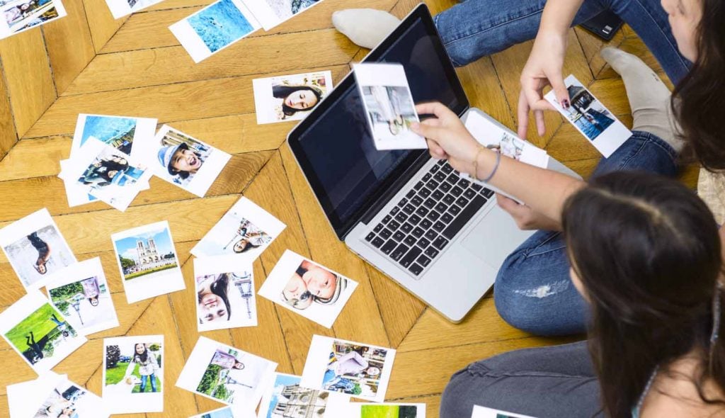 two girls looking at family photos with their laptop and photos scattered on hardwood floor