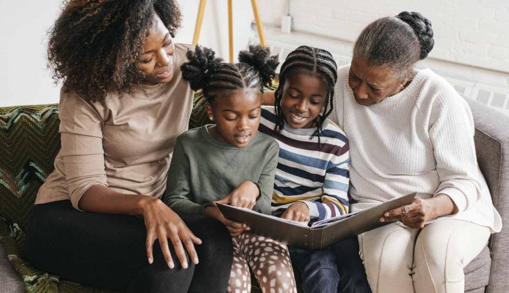 a mom, grandmother, and two young daughters sitting on a couch looking at family photos in an album