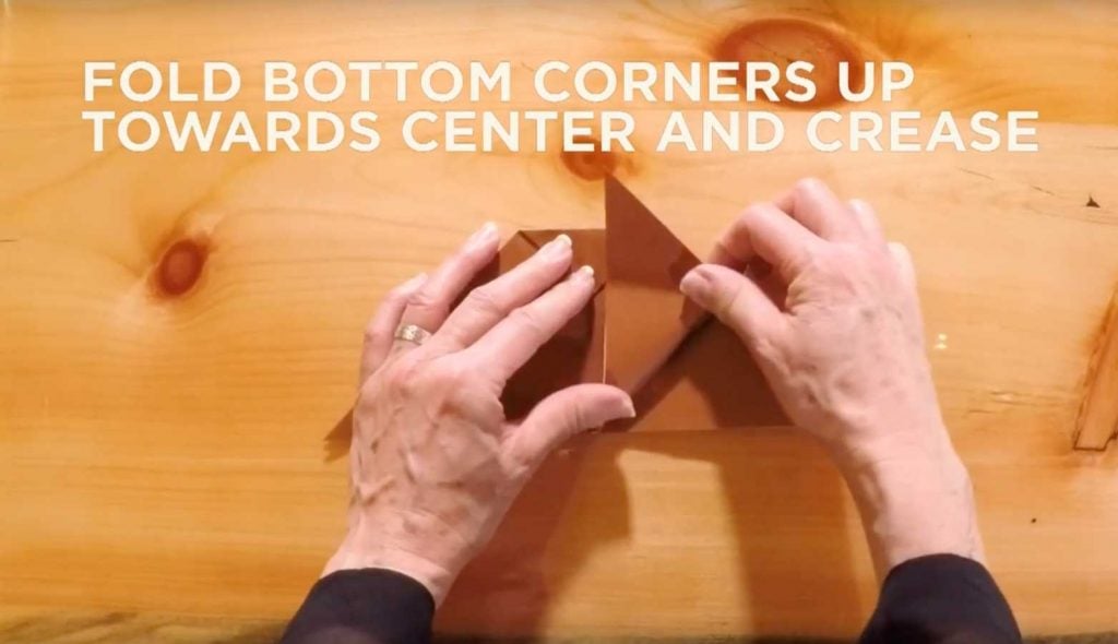 Paper being folded with text that says, "Fold bottom corners up towards center and crease."