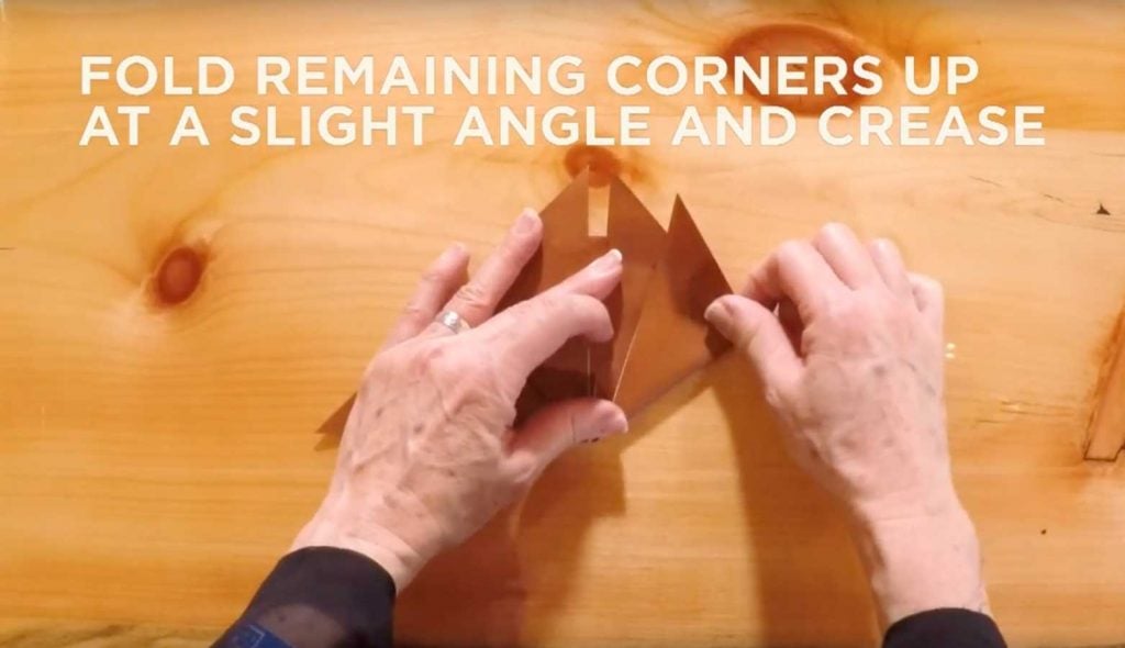 Paper being folded with text that says, "Fold remaining corners up at a slight angle and crease."
