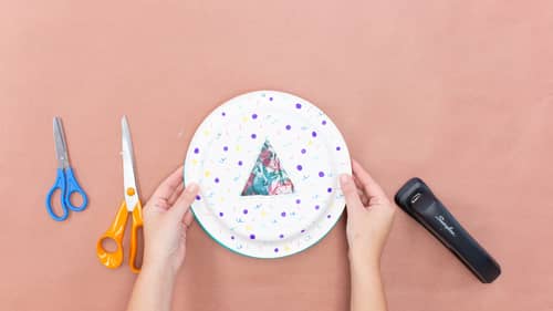 hands holding dotted paper plates, scissors and stapler