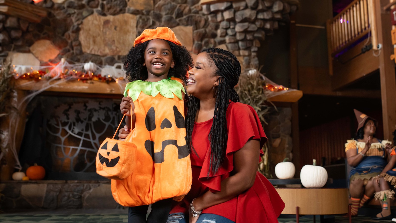 A smiling girl in a pumpkin costume holding a jack-o’-lantern treat bucket laughs beside a woman in a Halloween-decorated lodge.