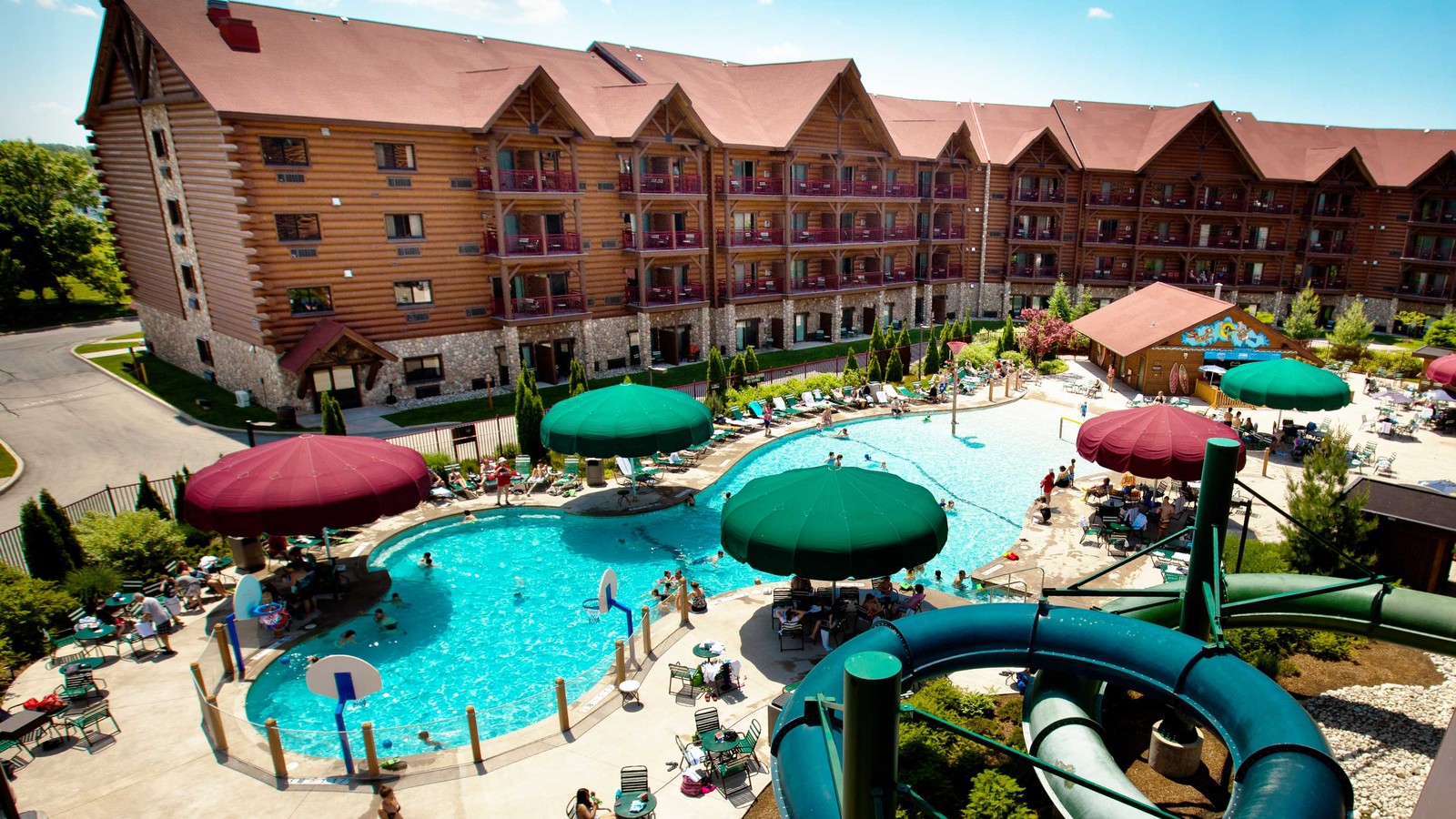 A large log cabin-style resort with a bustling outdoor pool area featuring water slides, shaded seating under colorful umbrellas, and guests enjoying a sunny day.  