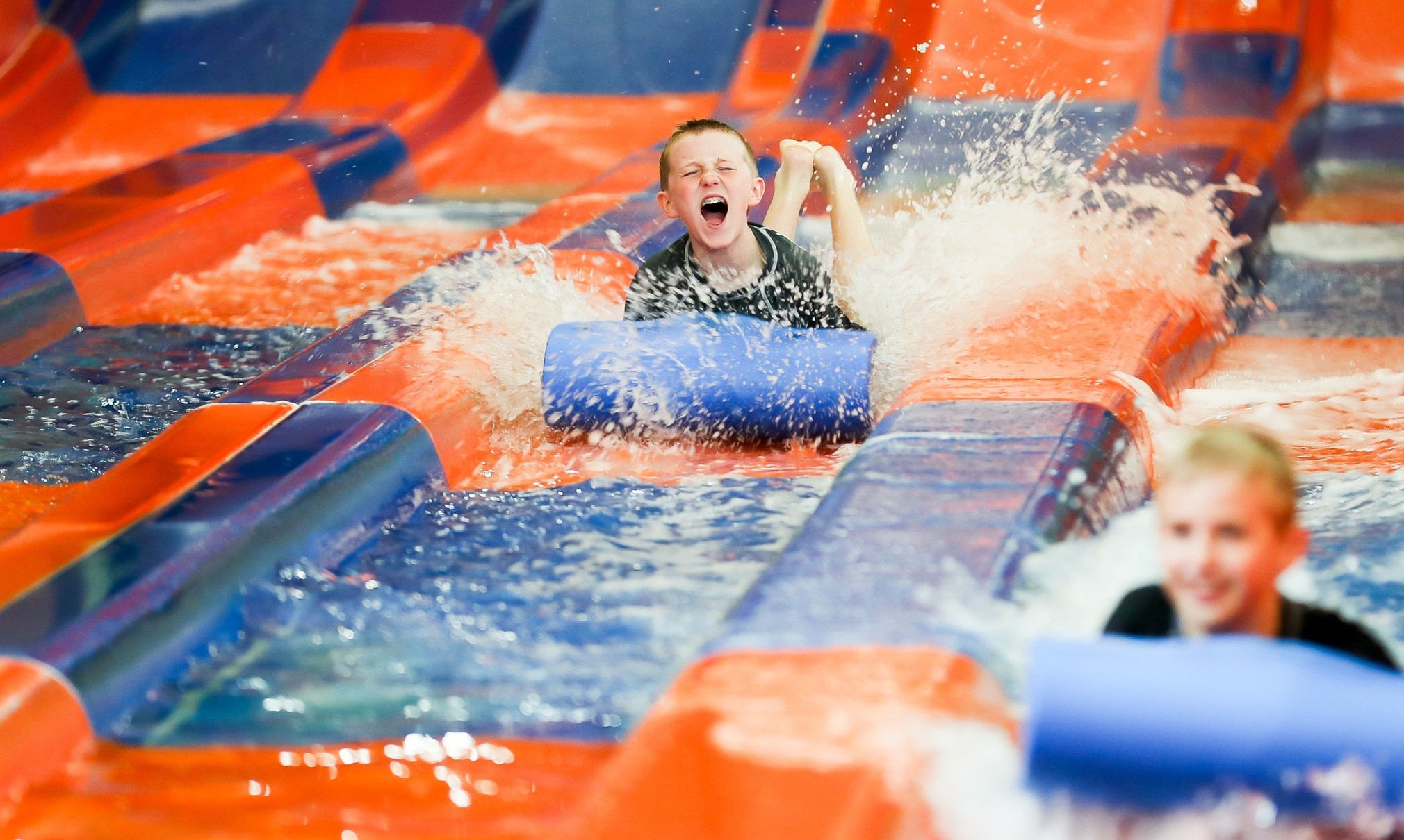 Two kids race down a waterslide laying on a blue mat