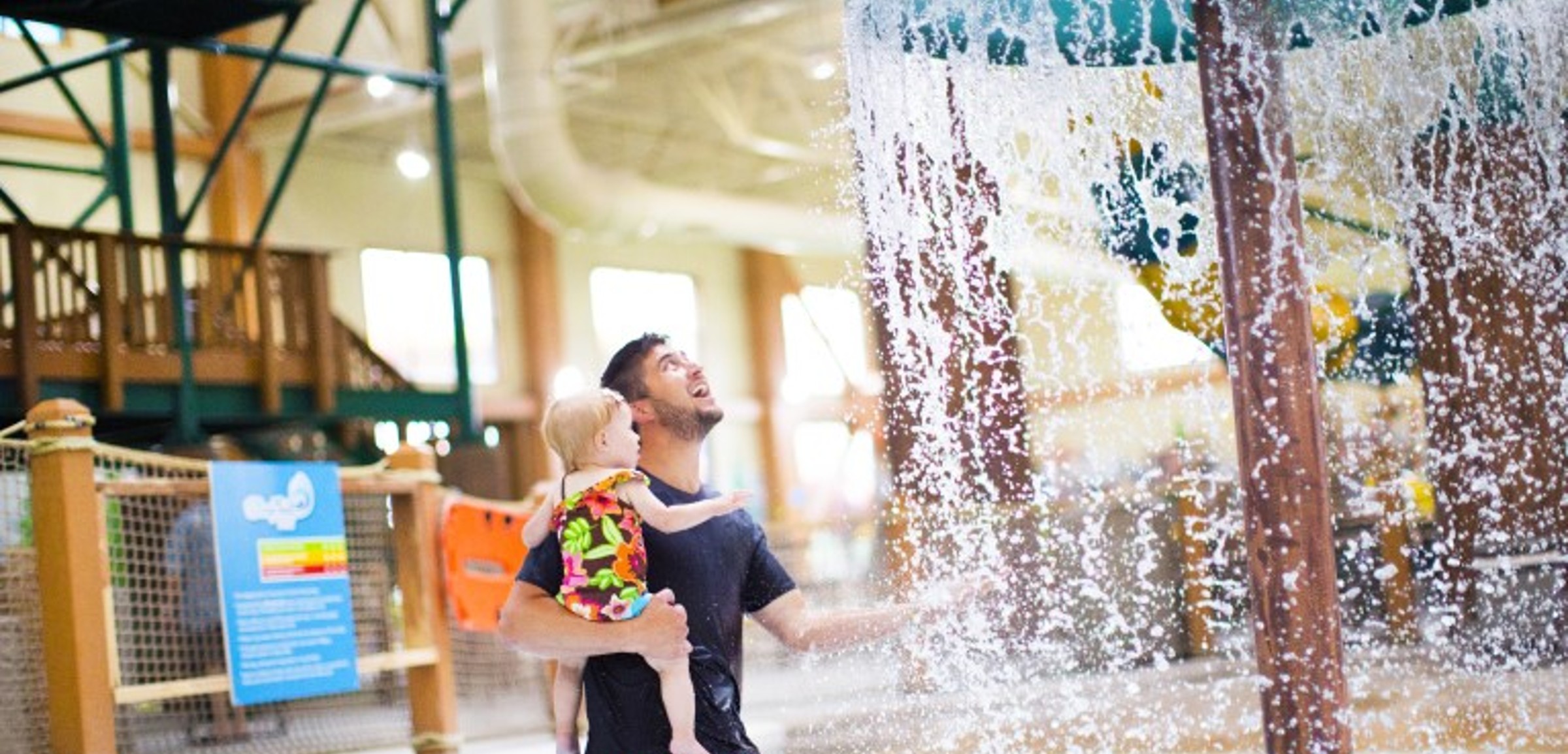 A father and toddler playing under a mushroom with water splashing 