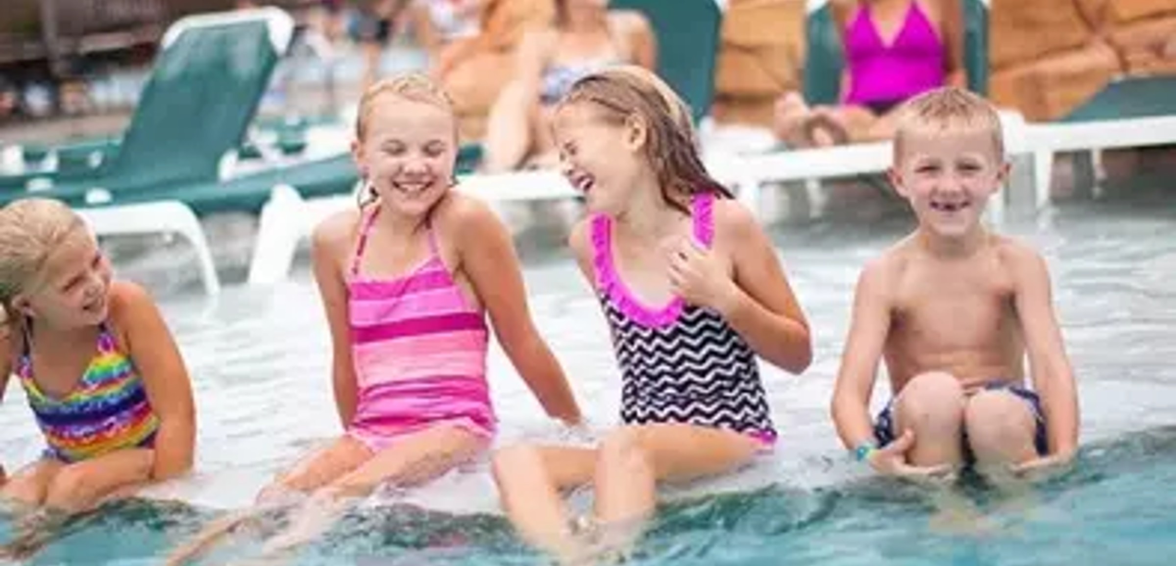 Four kids smile and laugh as they sit in a shallow outdoor pool 
