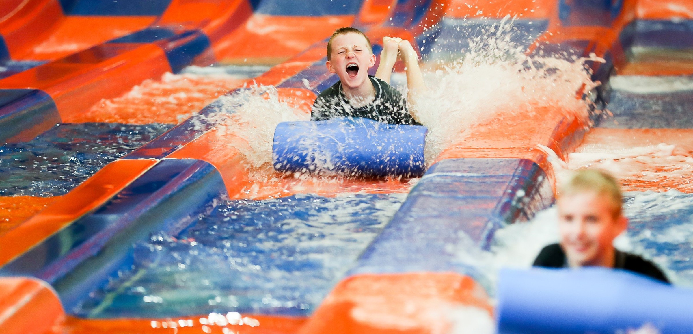Two kids race down a waterslide laying on a blue mat