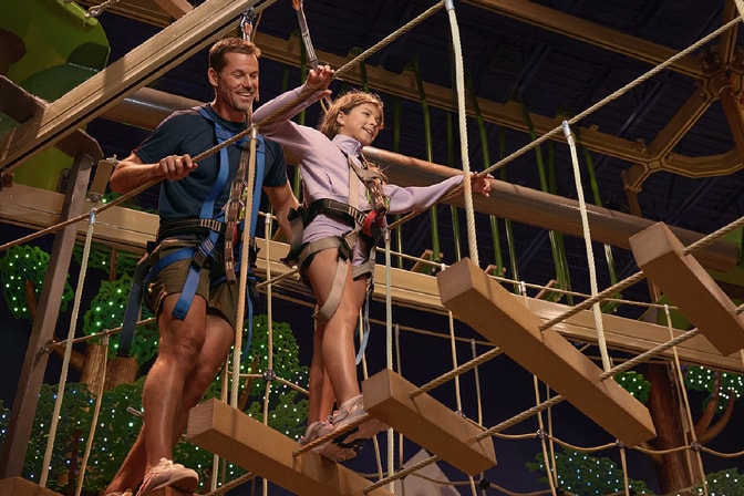 A man and a girl in harnesses smile as they cross a wooden rope bridge on an indoor ropes course.