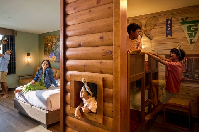 A family plays in a log-cabin-themed hotel suite as kids climb bunk beds and parents laugh by the bed.