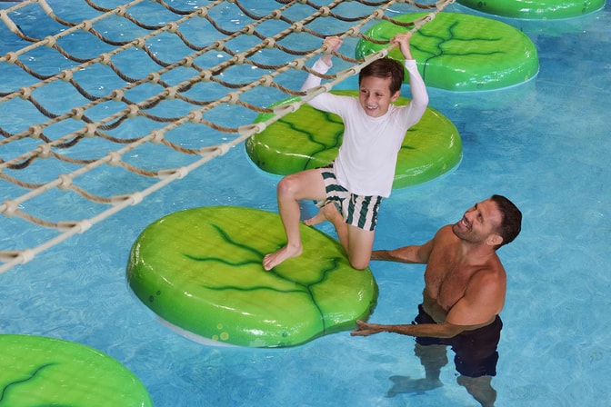 A smiling boy crosses floating lily pad stepping stones while holding a rope net as a man watches from the pool.