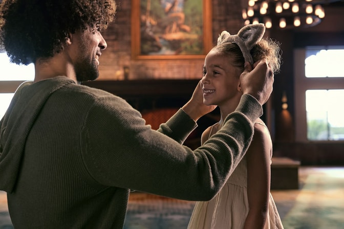 A man places plush wolf ears on a smiling girl in the rustic Great Wolf Lodge lobby, with a stone fireplace behind them.