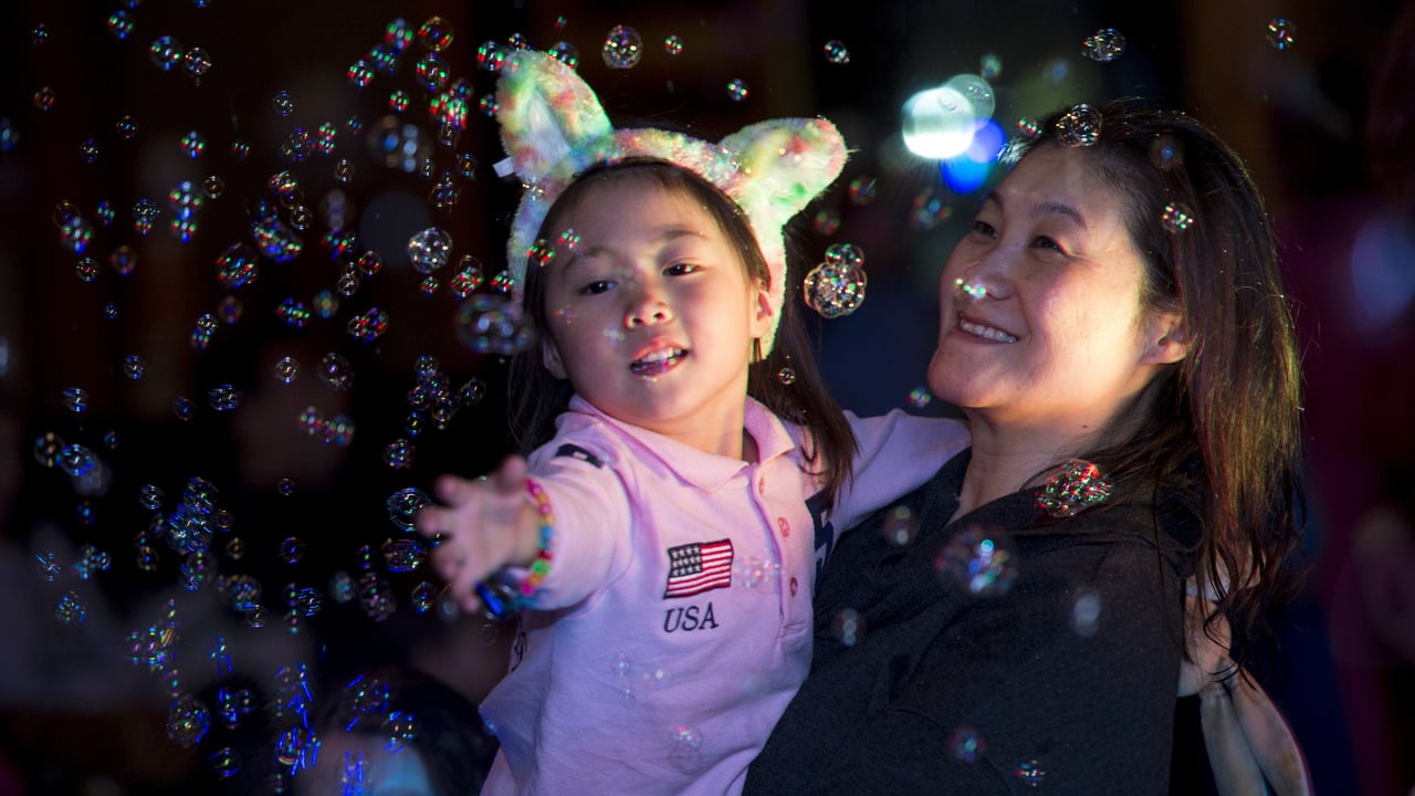 Smiling child with light-up ears reaching for bubbles while being held by a parent