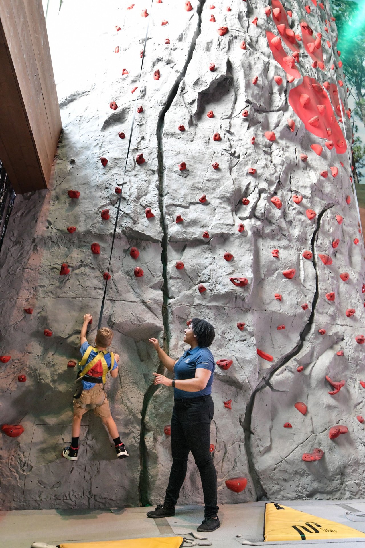 Child climbing indoor rock wall with instructor watching from below