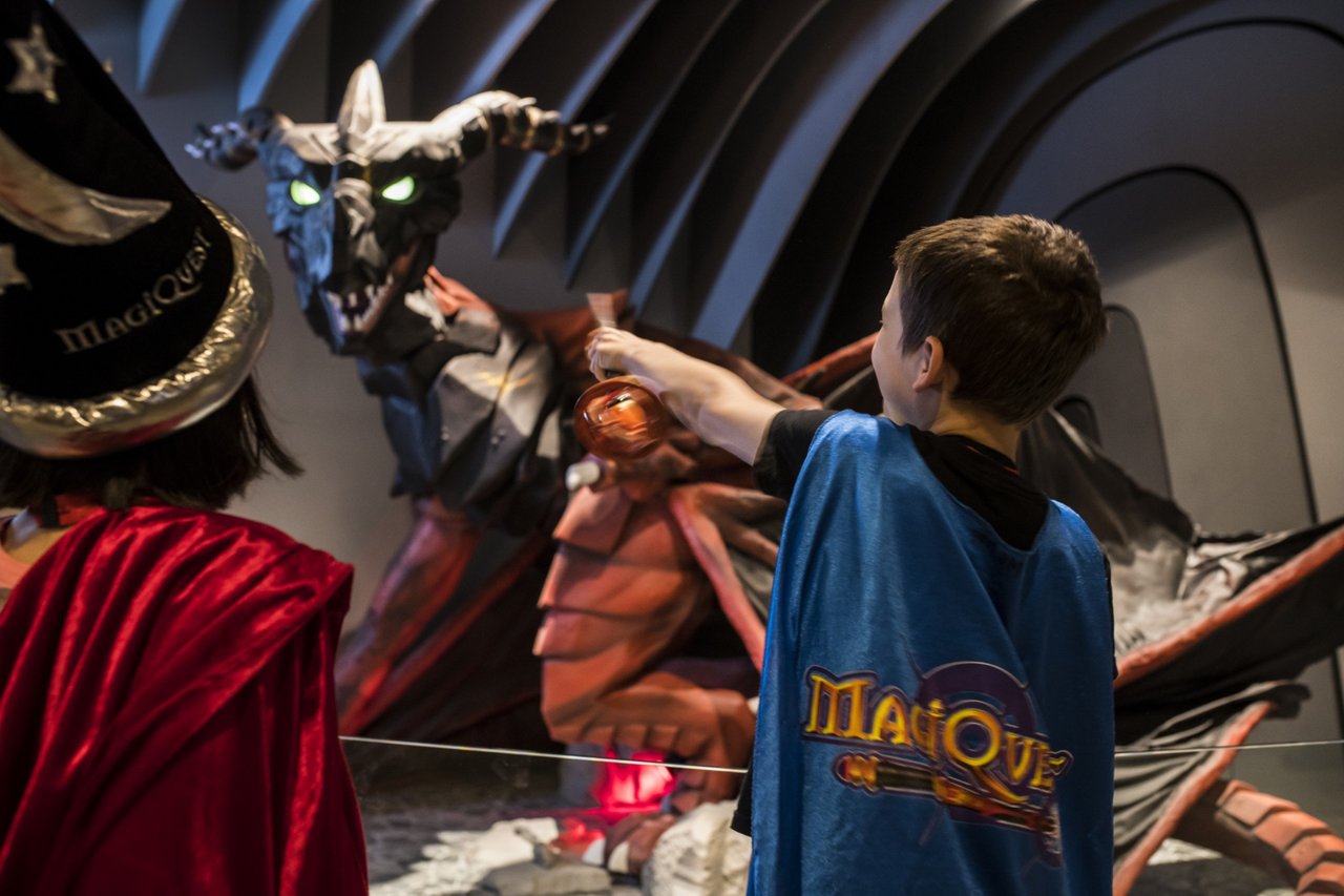 Two children wearing MagiQuest capes point their wands at MagiQuest dragon..