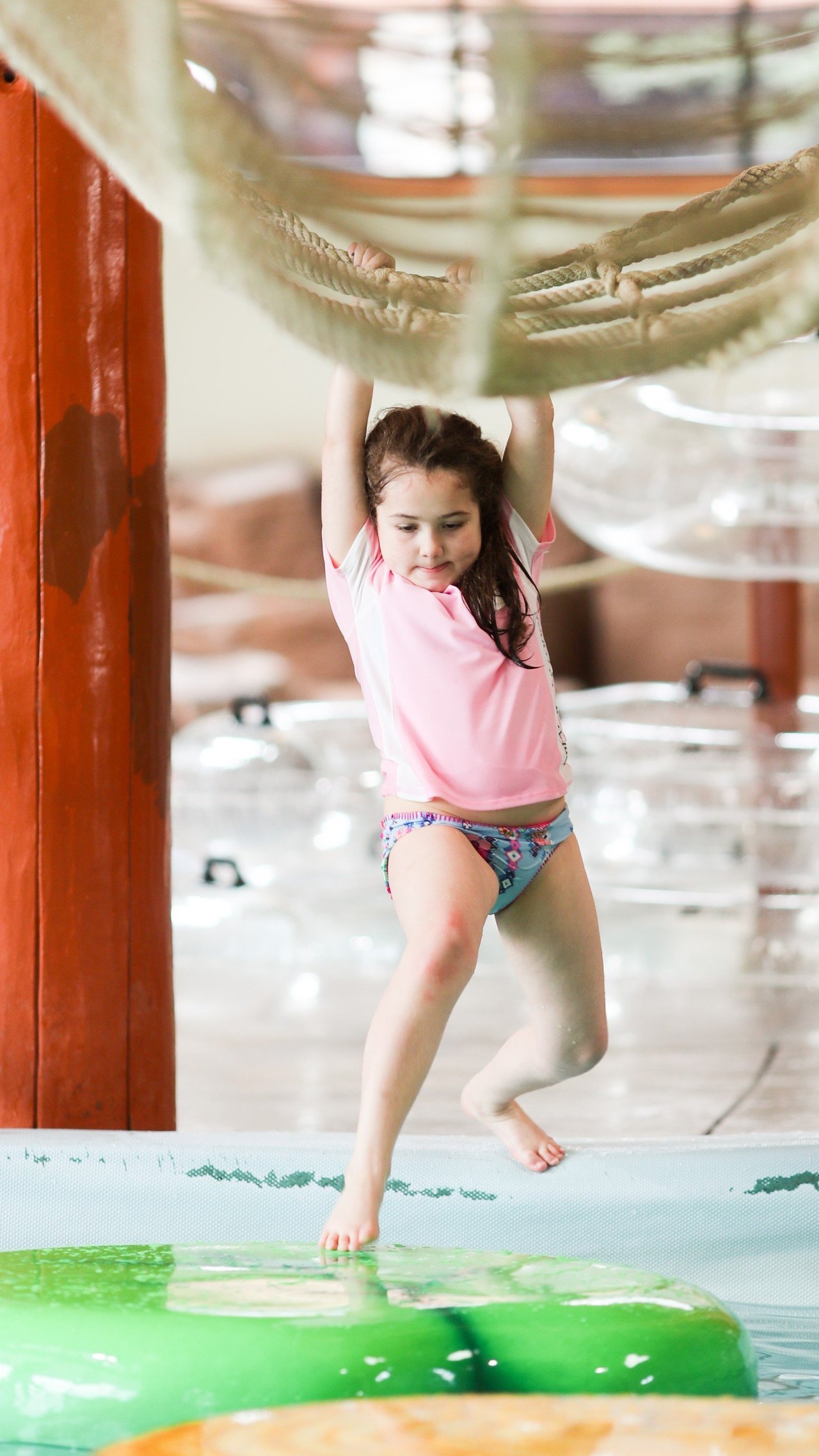 A young girl swinging from the rope bridge with her feet touching a lily pad pool float