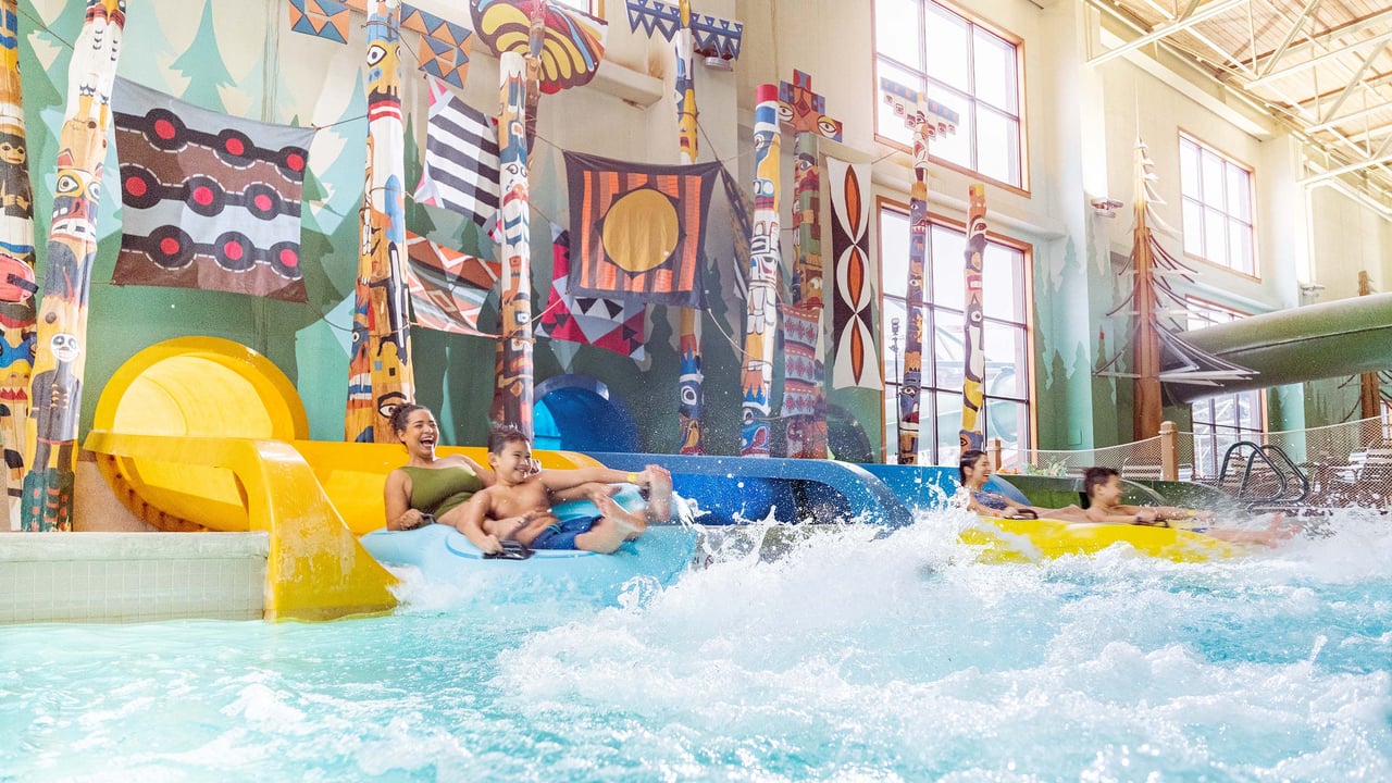 People exiting a yellow water slide into a splash pool, surrounded by colorful totem poles and Indigenous-inspired artwork in an indoor water park.