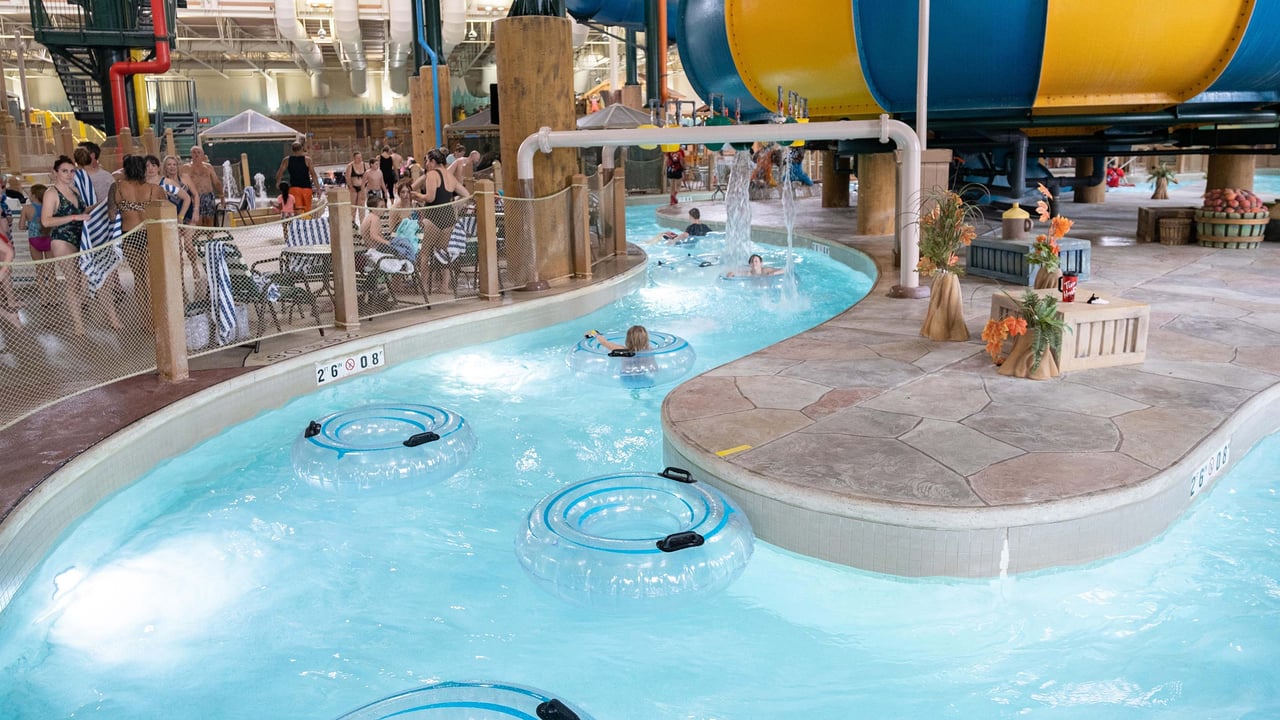 A group of people enjoy an indoor water park featuring a lazy river with clear inner tubes, cascading water features, and a large yellow-and-blue water slide in the background.