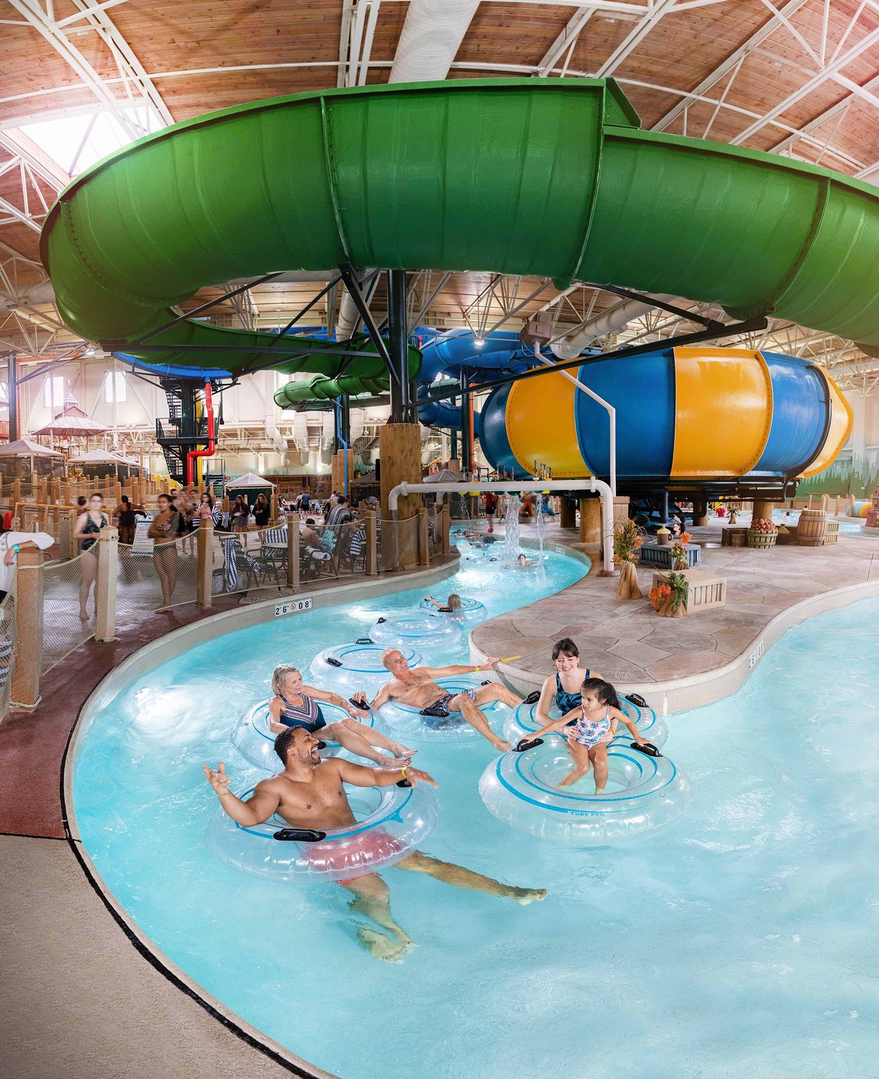 Children enjoy an indoor water park featuring a lazy river with clear inner tubes, cascading water features, and a large yellow-and-blue water slide in the background.
