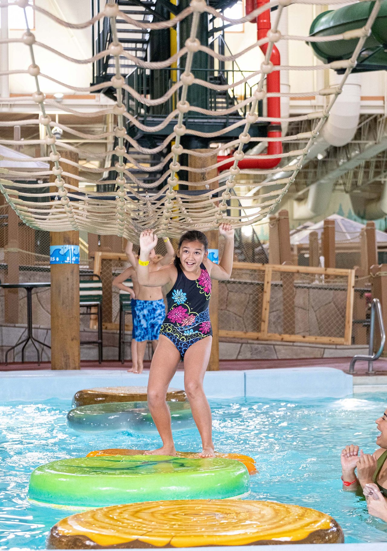 A young girl smiles as she grabs on to the rope net over a pool while balancing on inflatable lily rafts.