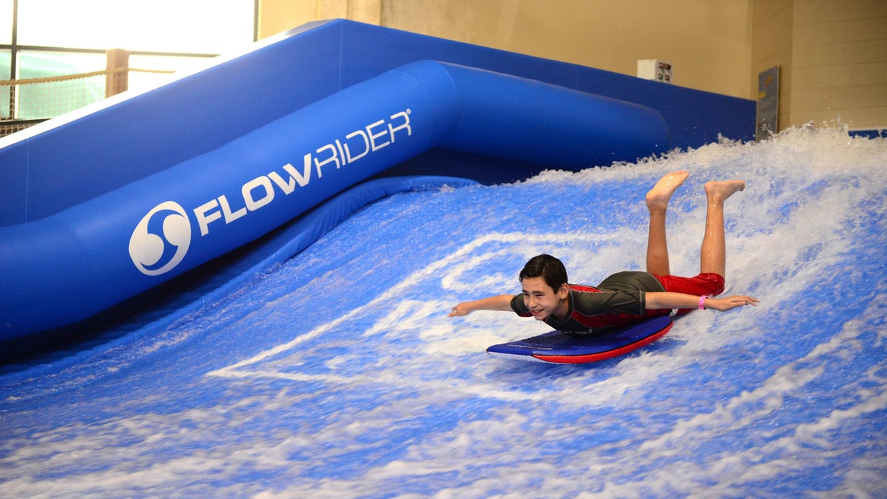 A boy laying on his belly on  blue boogie board going down the slide with his arms out and his feet up 