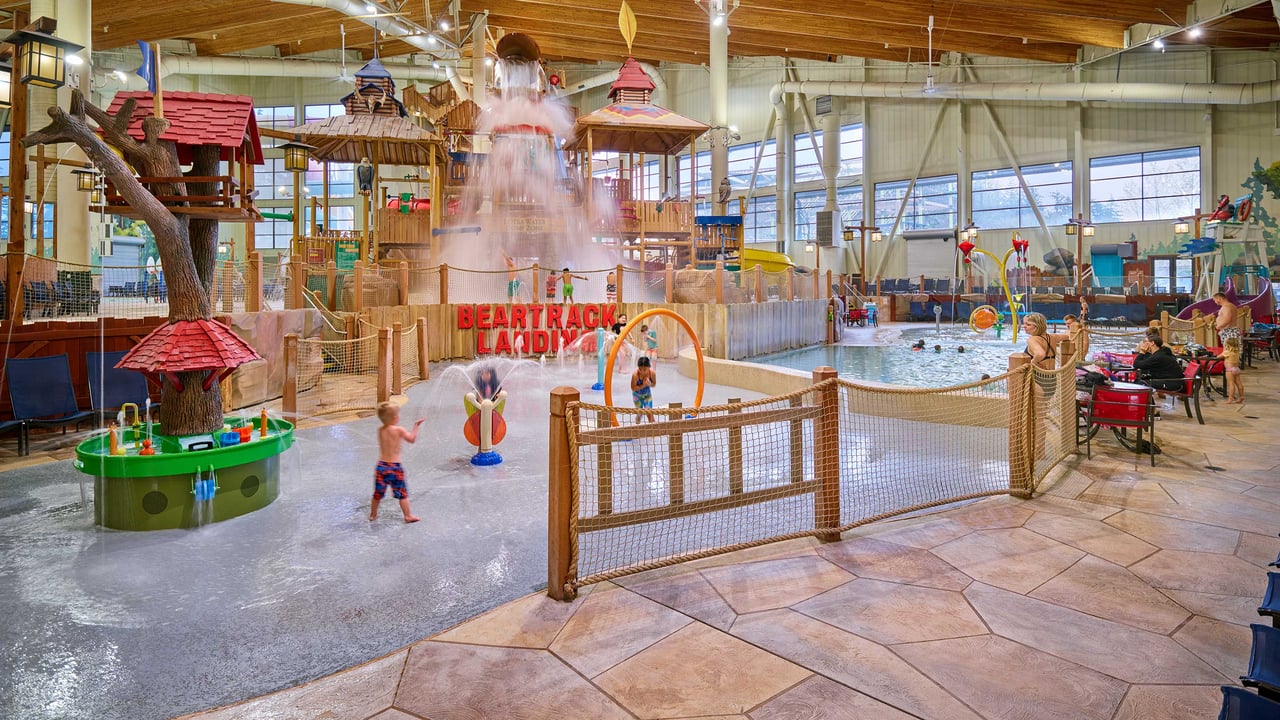 Kids are joyfully playing under a water feature at an indoor water park, surrounded by colorful play structures.