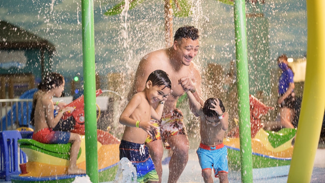 A man and two young boys are joyfully playing under a water feature at an indoor water park, surrounded by colorful play structures.