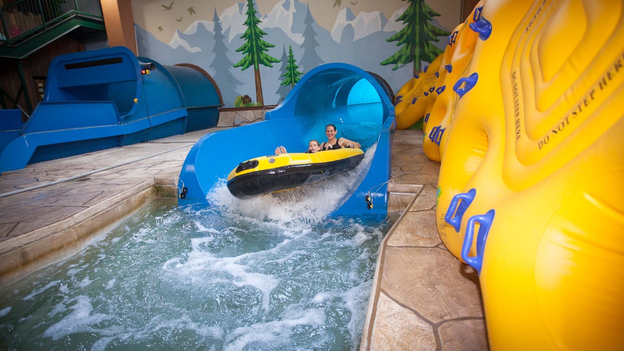 Two people in a yellow tandem raft landing in a pool of water at the end of a indoor water slide.