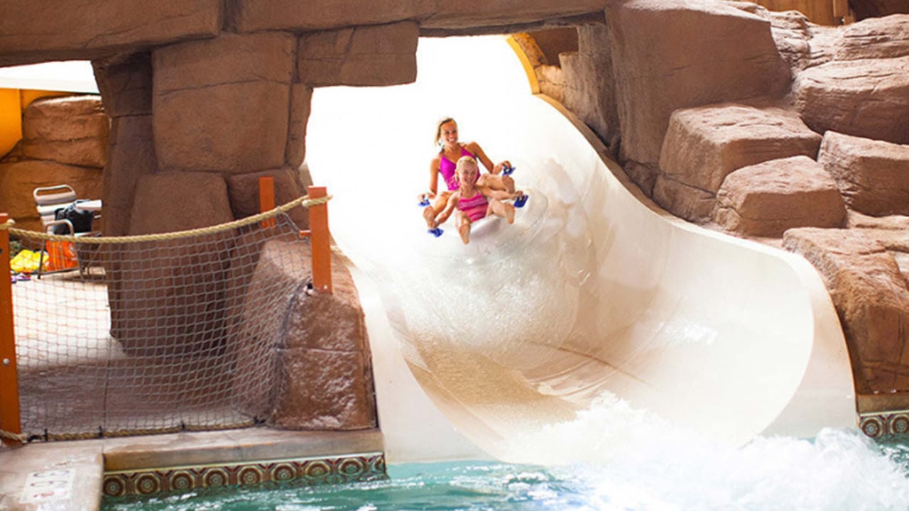 Woman and child in clear tubes riding down a white waterslide together surrounded by rocky decor