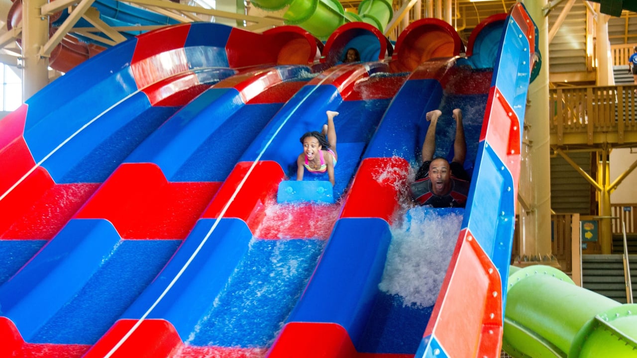 A young girl and her father race down a waterslide laying on a blue mats