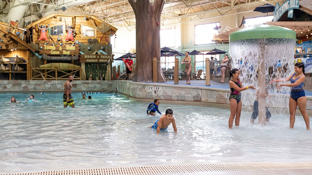 An indoor water park with a rustic theme featuring a shallow pool area where children and adults play, a central waterfall feature, a decorative large tree trunk, and a log cabin structure labeled "Tooth & Claw Snack Shop" in the background.