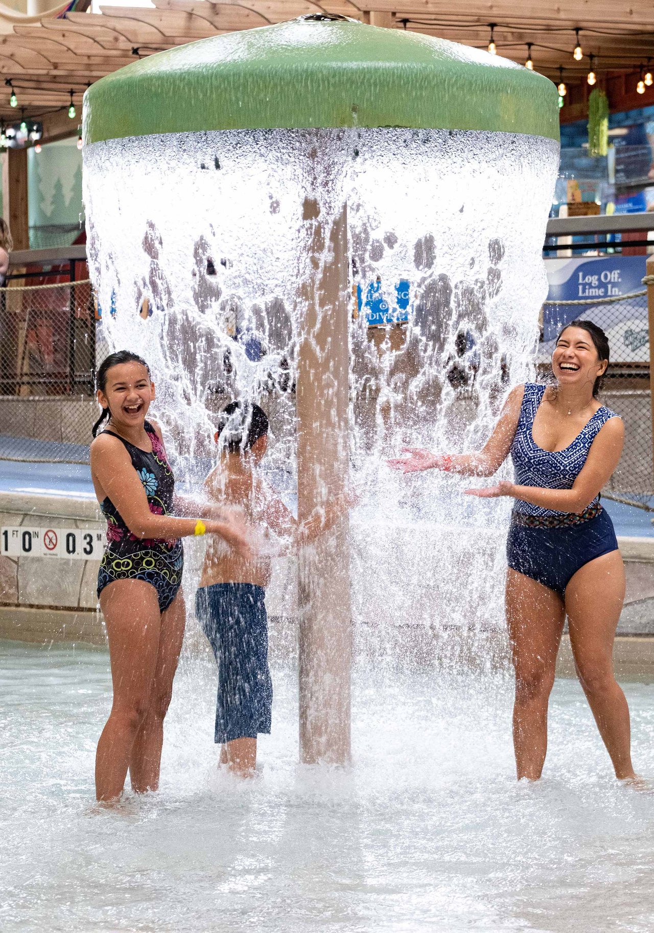 Two women in swimsuits laughing and playing in a water park feature with a large green-topped waterfall bucket that cascades water around them, while a child plays in the middle of the water stream.