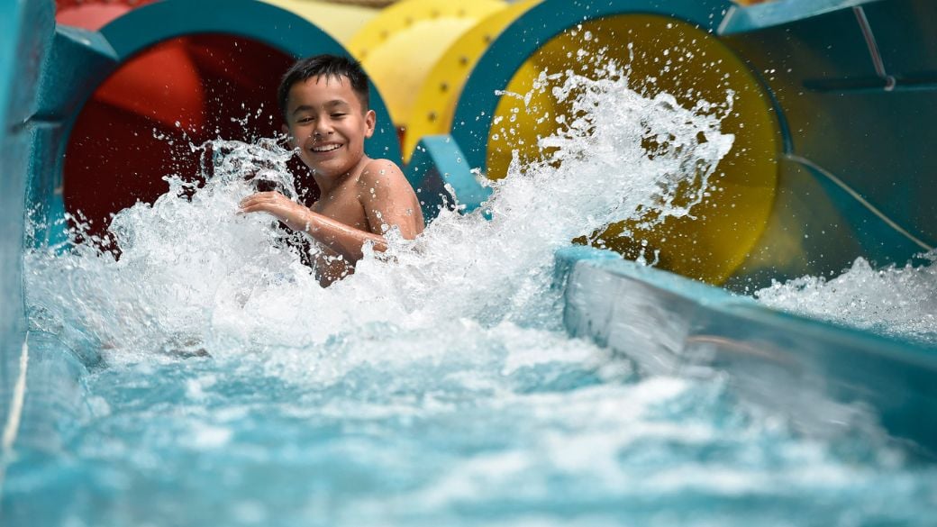 A young boy smiling as he splashes down a water slide into the landing pool.