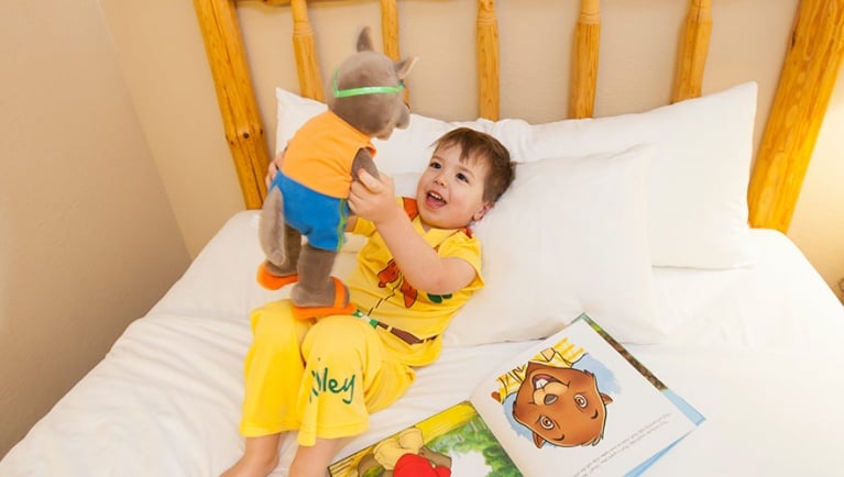 A boy lies in bed while holding up a stuffed animal available at Great Wolf Lodge indoor water park and resort.