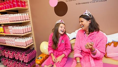 A mother and aughter sit on a banana split-shaped couch while waiting for service at Scooops Kid Spa at Great Wolf Lodge indoor water park and resort.