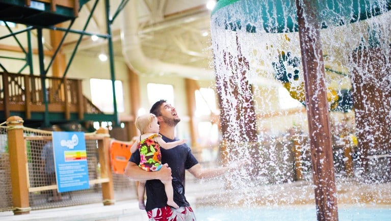 A father and toddler playing under a mushroom with water splashing 