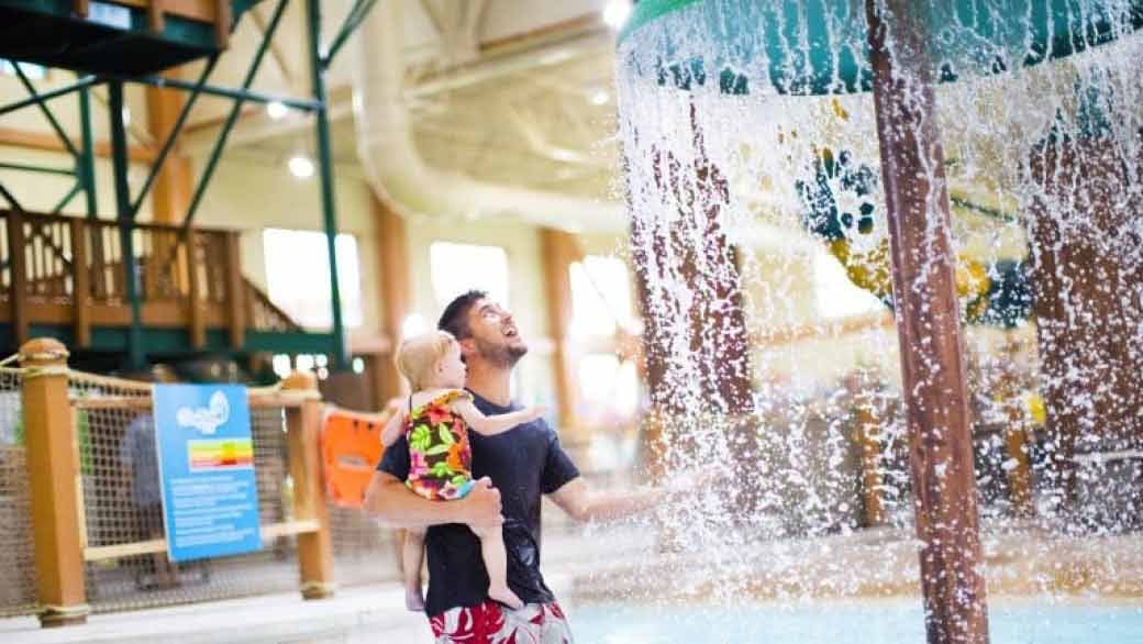 A father and toddler playing under a mushroom with water splashing 