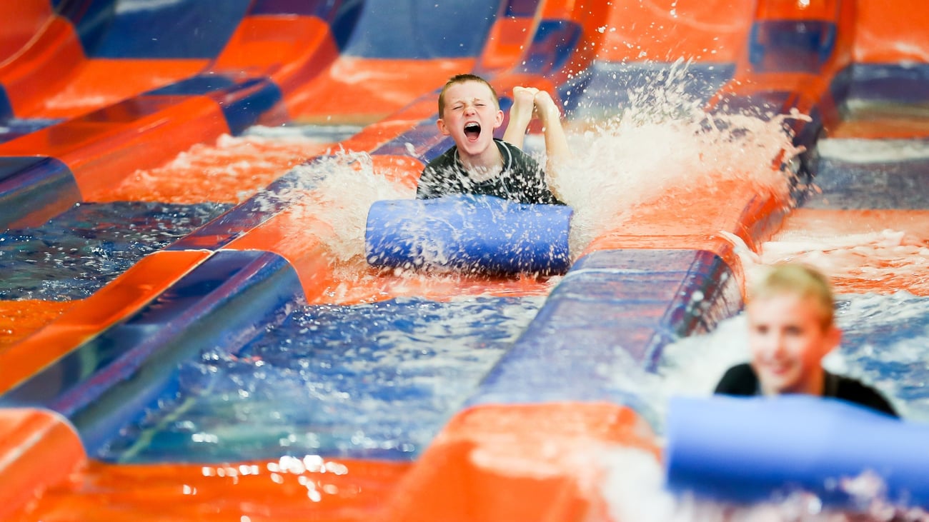 Two kids race down a waterslide laying on a blue mat