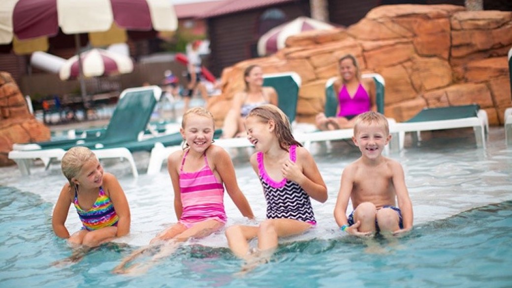 Four kids smile and laugh as they sit in a shallow outdoor pool 