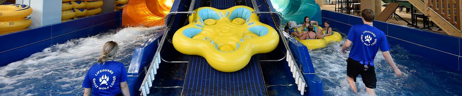 guests come down a water slide riding a yellow tube as pack members look at them for providing help