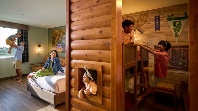 A family plays in a log-cabin-themed hotel suite as kids climb bunk beds and parents laugh by the bed.