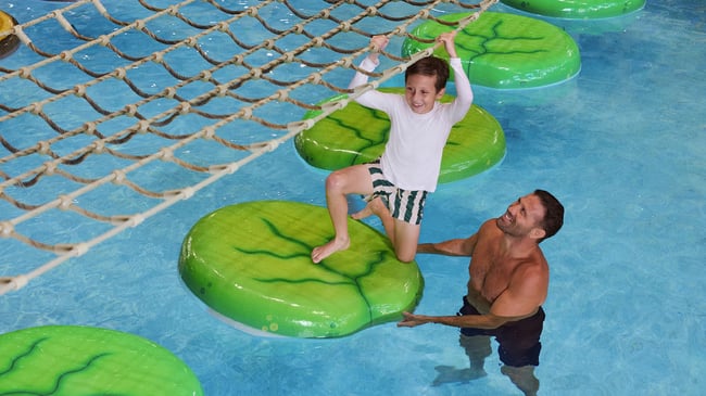 A smiling boy crosses floating lily pad stepping stones while holding a rope net as a man watches from the pool.