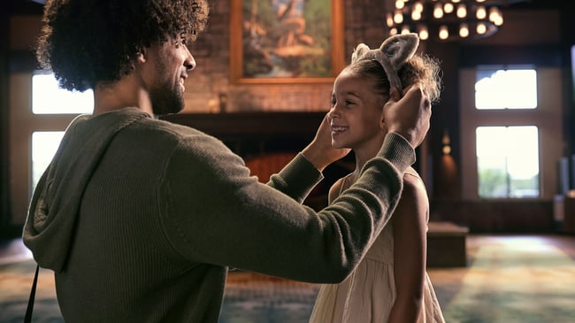 A man places plush wolf ears on a smiling girl in the rustic Great Wolf Lodge lobby, with a stone fireplace behind them.