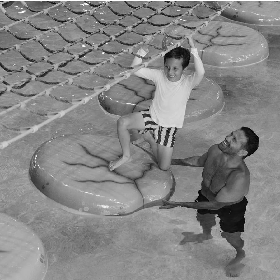 a girl  holding ropes passes over lily pads in pool as her father looks at her from the pool