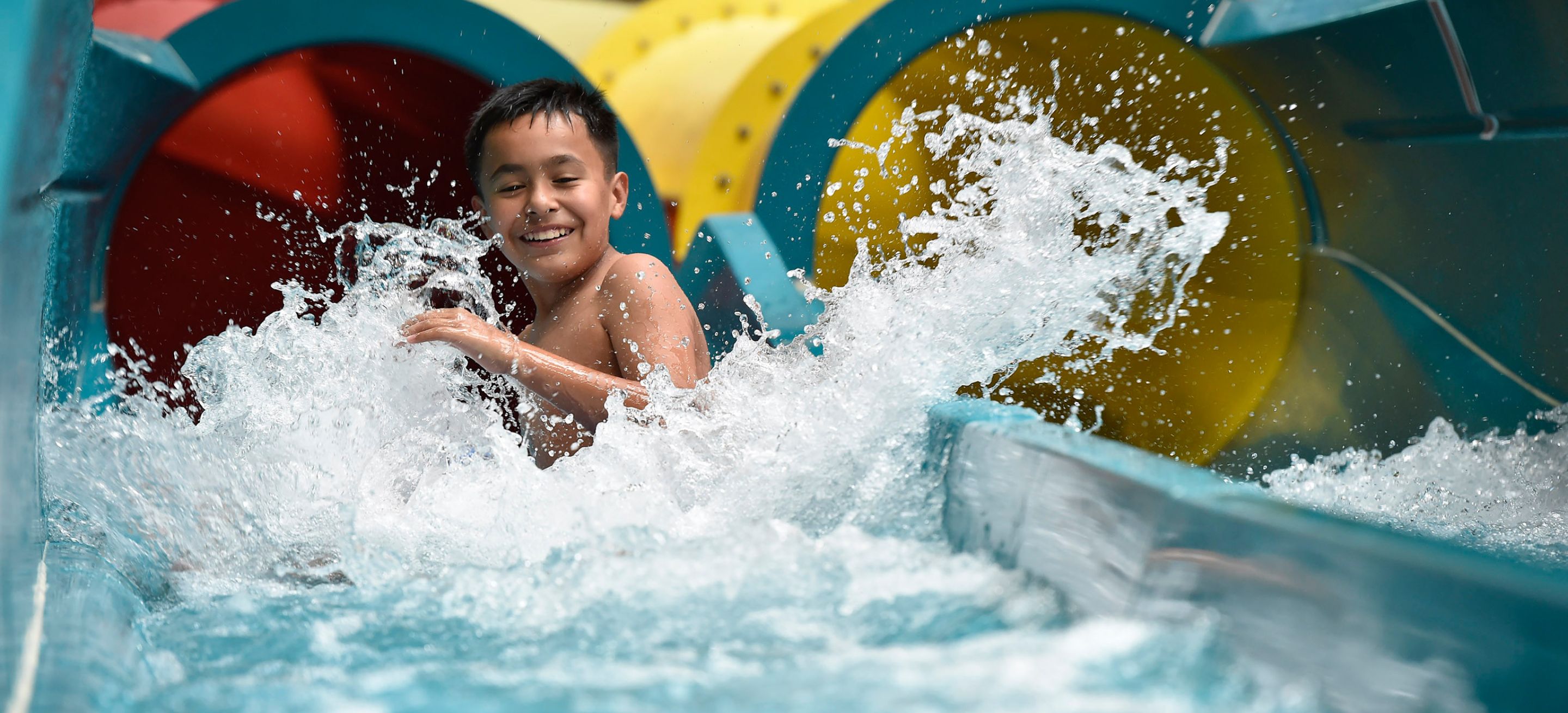 A young boy smiling as he splashes down a water slide into the landing pool.