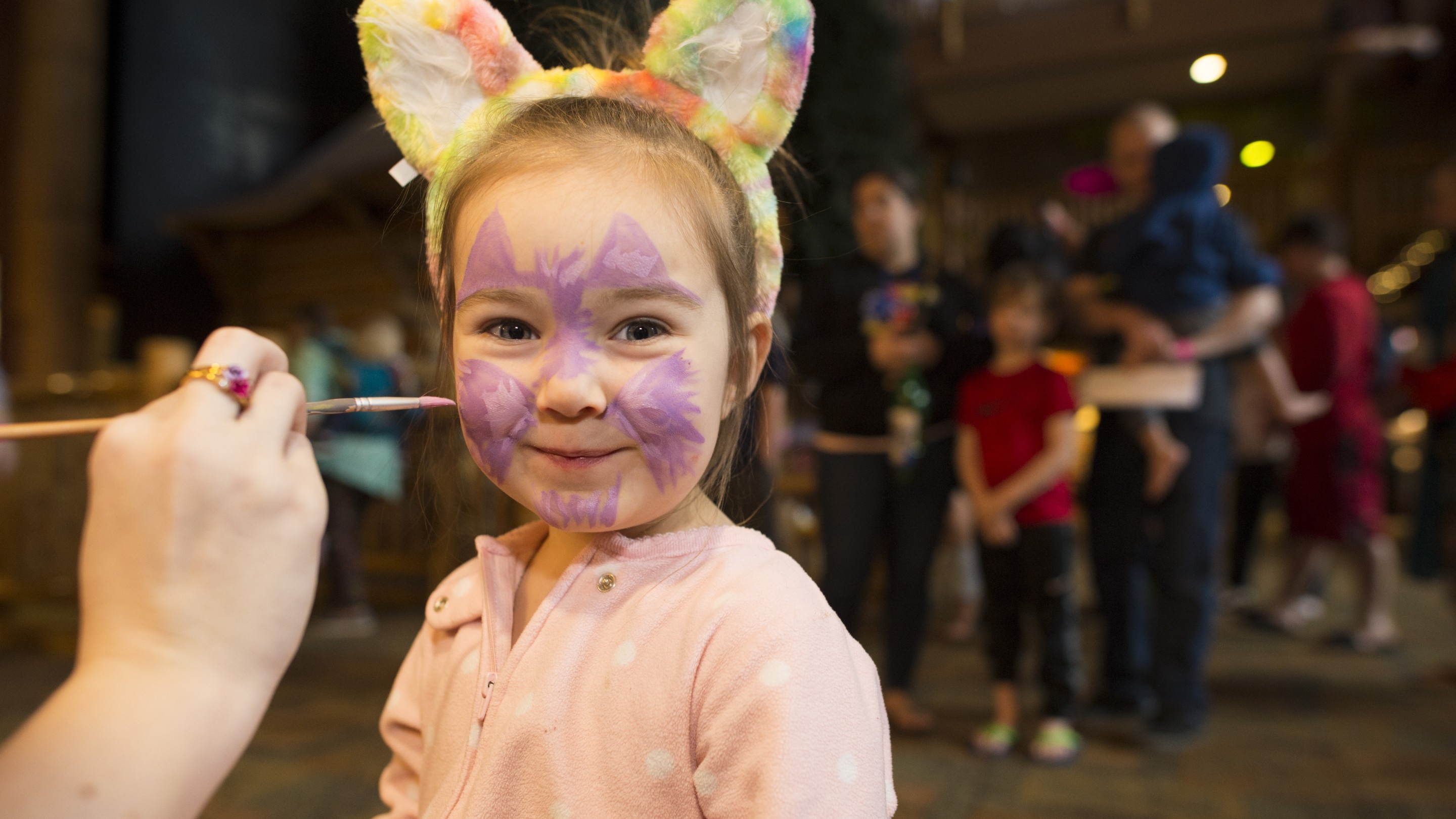 A little girl with colorful animal ears smiles while getting her face painted like a purple cat
