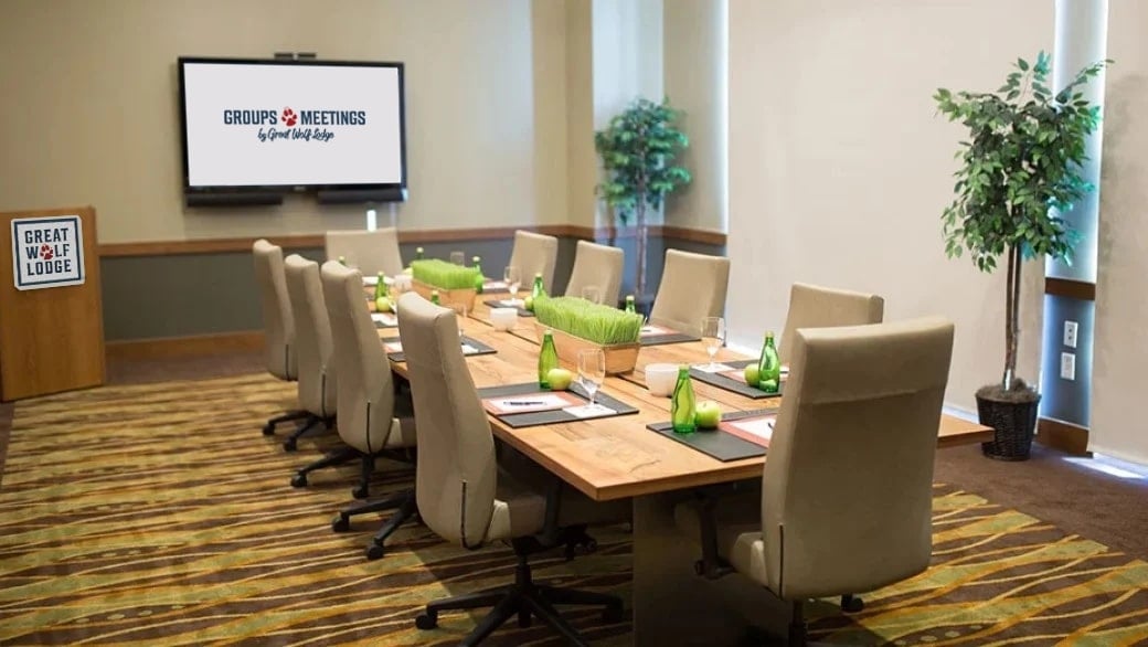 chair surround a long desk with water and green center pieces on it facing a wall mounted TV