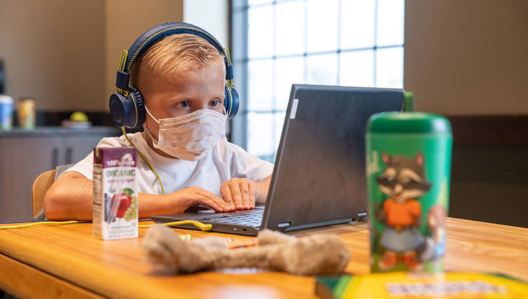 A child wearing a mask attends their daily lessons via laptop.