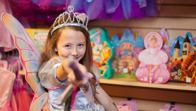 A girl wearing a tiara and fairy wings points at the camera while shopping 