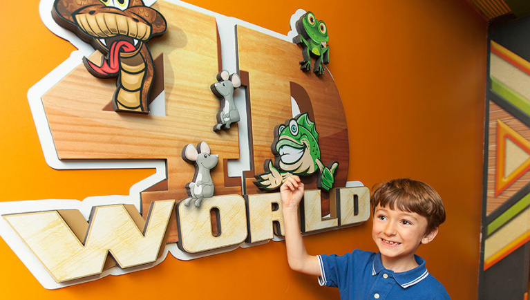A boy smiles next to the logo for 4D World at Great Wolf Lodge indoor water park and resort.