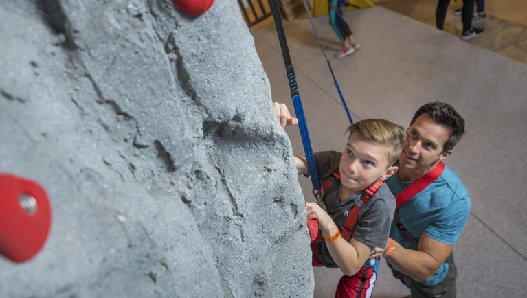 A father helps his son climb a rock wall ar Great Wolf Lodge indoor water park and resort indoor water park 